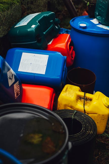 The image shows a collection of various plastic and metal containers, including a large blue drum, a yellow jerry can, a red fuel can, and a dark green toolbox, arranged outdoors on a grassy or earthy surface. Some of the containers have labels or stickers, and they are positioned close together, with a coiled black hose or cable partially visible at the bottom right corner. The background includes greenery, suggesting a garden or yard setting. The scene appears to depict materials commonly associated with waste or bulk rubbish, possibly prepared for collection or disposal, aligning with services offered by rubbish removal specialists such as Rubbish Junk, who handle on-site clearance or alternative private waste disposal solutions, especially for bulky or hazardous items.